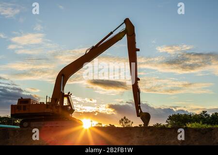 Silhouette eines großen Raupenbaggers, der den Boden vor Ort gräbt. Aufnahmen bei Sonnenuntergang mit Sonnenlicht und Flare Effekt Stockfoto