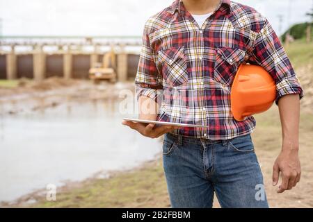 Junger asiatischer Ingenieur arbeitet vor Ort am Staudamm und kontrolliert, während der Bagger die Erde gräbt Stockfoto