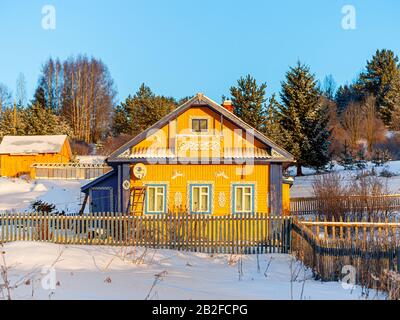 Winterblick auf Holzhaus im Dorf im russischen Norden. Altes Haus auf dem Land in wunderschöner Sonneneinstrahlung gegen einen blauen Himmel. Kaltes Wetter Stockfoto