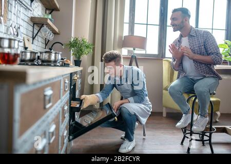Junger, fadenhaariger Mann, der Gebäck aus dem Ofen nimmt, ein weiterer sitzt daneben. Stockfoto