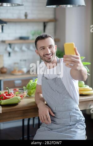 Junger Kerl in einer Schürze, der ein selfie in der Küche nimmt. Stockfoto