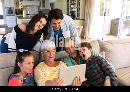 Die Vorderansicht von sechs Mitgliedern einer kaukasischen Mehrfamilien-Familie zu Hause auf einem Sofa im Wohnzimmer, versammelt um die Großmutter, die hält Stockfoto