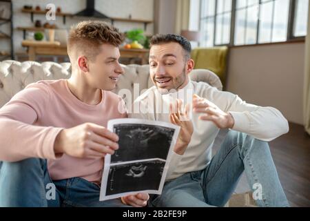 Männliches Paar mit Ultraschallbildern, die zu Hause auf dem Boden sitzen. Stockfoto