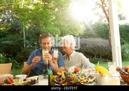 Vorderansicht eines älteren kaukasischen Ehepaares, das draußen an einem Esstisch sitzt, das für eine Mahlzeit steht, mit den Armen verbunden und lächelnd trinkt. Familie genießen Stockfoto