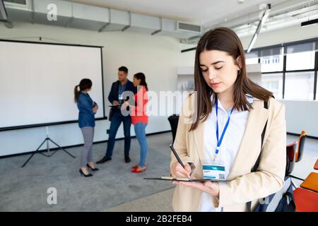 Vorderansicht einer kaukasischen Geschäftsfrau, die in einem modernen Büro arbeitet und in einer Zwischenablage schreibt, mit Kollegen neben einem Whiteboard, das miteinander spricht Stockfoto