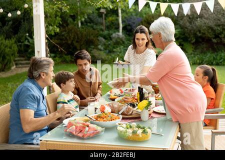 Vorderansicht einer mehrköpfigen kaukasischen Familie, die draußen an einem Esstisch sitzt, für eine Mahlzeit, Unterhaltung und Essen. Familie, die Zeit genießt Stockfoto