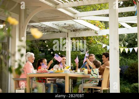 Niedriger Winkel Vorderansicht einer mehrköpfigen kaukasischen Familie, die draußen an einem Esstisch sitzt, der für eine Mahlzeit eingerichtet ist, US-Flaggen feiert und schwenkt. Familie Stockfoto