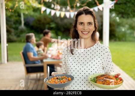 Porträt einer lächelnden kaukasischen Frau, die Teller mit Essen hält, mit ihrer mehrköpfigen kaukasischen Familie, die draußen an einem Esstisch sitzt Stockfoto