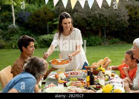 Vorderansicht einer mehrköpfigen kaukasischen Familie, die draußen an einem Esstisch sitzt, für eine Mahlzeit, Unterhaltung und Essen. Familie, die Zeit genießt Stockfoto