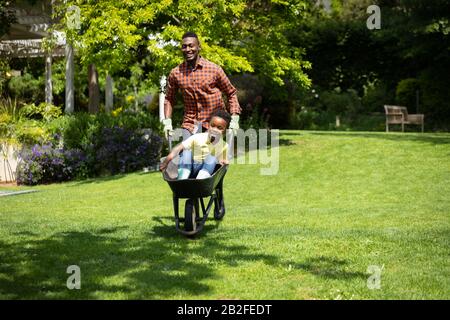 Vorderansicht eines glücklichen afroamerikanischen Mannes im Garten, der seinen Sohn in einer Schubkarre trägt und dabei mit einem Lächeln vor die Kamera lächelt. Familie genießt Zeit zu Hause, Lifestyle Stockfoto