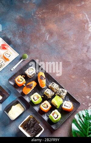 Frisch gekochtes Sushi-Brötchen-Menü auf einer Keramikplatte mit weißer Soße, Nori-Stücke auf einem dunklen Betontisch und grünem immergrünen Blatt, Kopierraum. Japane Stockfoto