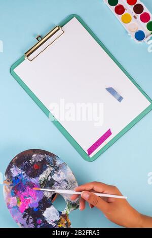 Kunsthandwerk von Malmaterial. Hand der Frau mit Pinsel über Aquarellfarben, Künstlerpalette und leerem Blatt Papier auf hellblauem Hintergrund, Stockfoto