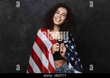 Bild einer jungen netten Frau mit einem Tattoo, das mit der Kamera lächelt und die amerikanische Flagge über schwarzem Hintergrund isoliert hält Stockfoto