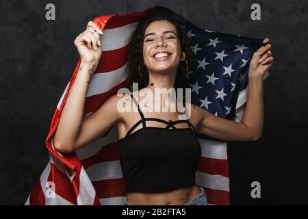 Bild einer jungen kaukasischen Frau mit einem Tattoo, das mit der Kamera lächelt und die amerikanische Flagge über schwarzem Hintergrund isoliert hält Stockfoto