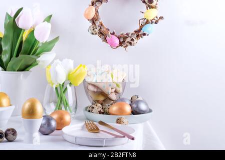 Feiertag Ostertischeinstellung mit handgemalten Eiern, gebackenen Plätzchen und frischen Blumen auf einem Tisch mit weißem Tuch. Stockfoto