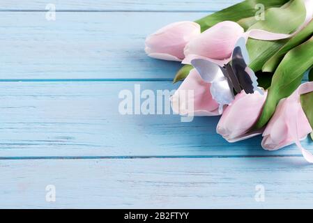 Grußkarte mit frischen, zarten, rosafarbenen Tulpen und Schmetterling aus Kunsthandwerk auf pastellblauem Holzhintergrund. Stockfoto