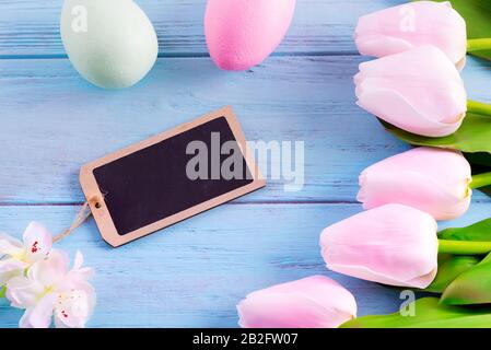 Ferienkarte mit frischen, zarten Tulpenrahmen und Schwarzen Brett für Nachrichten auf pastellblauem Holzhintergrund. Stockfoto