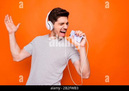 Foto von coolem Kerl fröhliche Party-Stimmung kühlende Hör-Ohrhörer halten Telefon wie Mikrofonaufnahme Single Wear gestreiftes T-Shirt isoliert