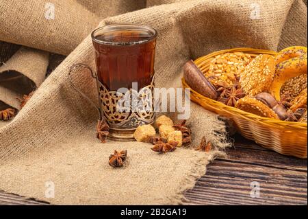 Ein Glas Tee in einem Vintage-Cup-Halter und Plätzchen, Schokoladenkuchen und Bagels in einem Korbkorb auf dem Hintergrund rauer Homespun-Qualität. Nahaufnahme Stockfoto