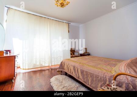 Ein großes Schlafzimmer mit Mahagoni, Korbstühlen und einem eleganten Schminktisch mit einem halbkreisförmigen Spiegel. Das Bett hat eine beige Decke in Quadraten. Stockfoto