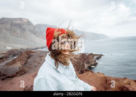 Lifestyle-Porträt einer stilvollen Frau, die bei starkem Wind eine Reise an einer felsigen Meeresküste genießt. Reisen auf der Insel Tenera, Spanien Stockfoto