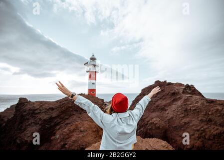 Unbeschwerte, stylische Frau, mit Jeans und rotem Hut gekleidet, die einen Ausflug auf einem felsigen Meeresufer in der Nähe des Leuchtturms macht und auf Nordwest-Insel auf der Insel Tenera, Spanien, unterwegs ist Stockfoto