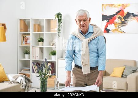 Portrait eines gutaussehenden Seniors in stilvollem Outfit, das am Tisch steht und die Papiere im Wohnzimmer betrachtet Stockfoto