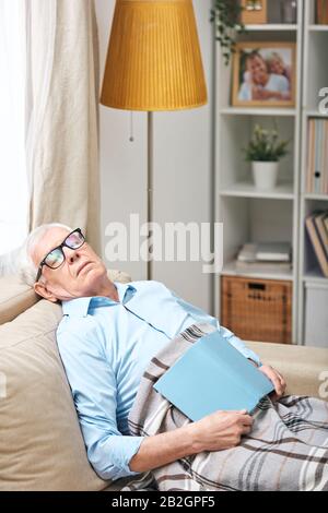 Der müde ältere Mann in einer Brille, die unter einem Sofa sitzt und mit Buch im Wohnzimmer schläft Stockfoto