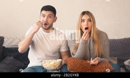 Überraschte das junge Paar mit Popcorn, das im Wohnzimmer Fernsehen sah Stockfoto