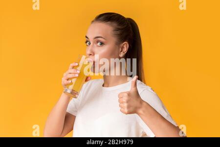 Durstige junge Frau trinkt frischen Orangensaft und stinkt den Daumen hoch Stockfoto