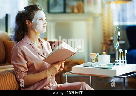 Junge Frau im Schlafanzug mit weißer Gesichtsmaske im Gesicht mit Buch und Blick in die Ferne im modernen Wohnzimmer am sonnigen Wintertag. Stockfoto