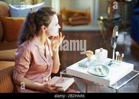 Stilvolle Frau im Schlafanzug mit weißer Gesichtsmaske im Gesicht und buchen Sie im modernen Wohnzimmer im sonnigen Wintertag. Stockfoto