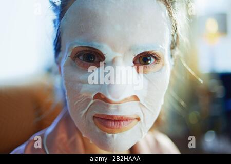 Porträt der eleganten 40 Jahre alten Frau im Schlafanzug mit weißer Gesichtsmaske im Gesicht im modernen Zuhause am sonnigen Wintertag. Stockfoto