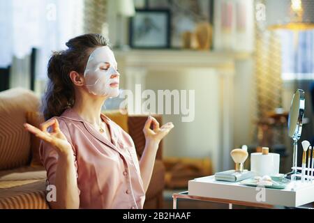 Entspannte, elegante Frau im Schlafanzug mit weißer Gesichtsmaske im Gesicht meditierend im modernen Haus am sonnigen Wintertag. Stockfoto