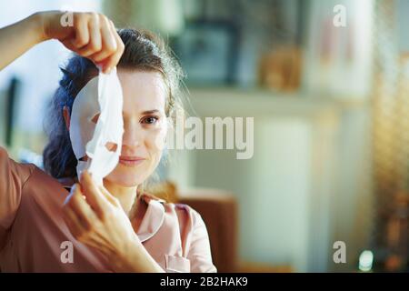 Porträt der modernen 40 Jahre alten Hausfrau im Schlafanzug im modernen Haus am sonnigen Wintertag weiße Gesichtsmaske entfernen. Stockfoto