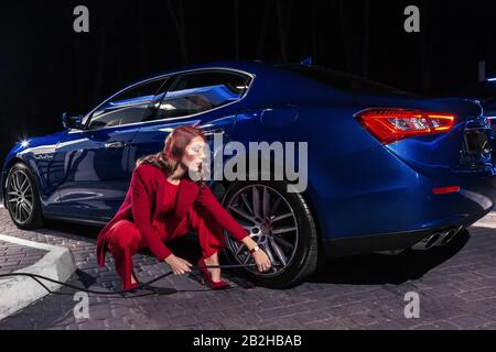 Die elegante blonde schöne Frau füllt Luft in einen Autoreifen, um den Druck des Autoreifens zu erhöhen. Schöne junge Frau im roten Anzug mit blauem Luxusauto Stockfoto