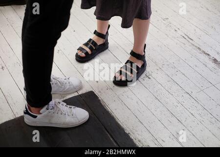 Zwei Paar Schuhe und Beine, weiße adidas Läufer und schwarze Jeans, Sandalen mit breiten Riemen und auf weißem Holzboden als Stillleben-Trend Stockfoto