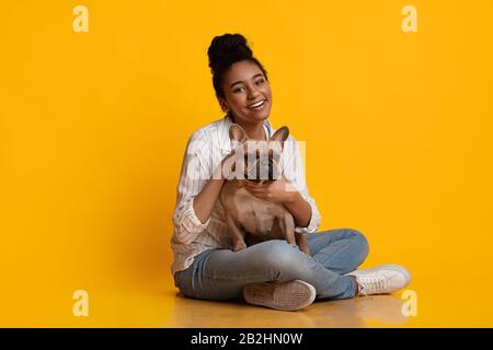 Fröhliches Afro Girl, Das Ihren Französischen Bulldog Puppy Umarmt Und Im Studio Posiert Stockfoto