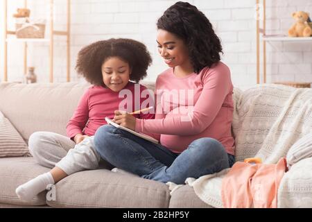 Die Schwangere Afro Mum Und Ihre Tochter, Die Sich Notizen Machen Und Babynamen Wählen Stockfoto