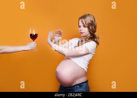 Schwangere Frau weigert sich, Wein zu trinken, und macht auf gelbem Hintergrund eine Stopp-Geste zum Glas Stockfoto