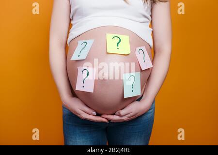 Schwangere Frau mit Fragezeichen auf Papieraufklebern auf Bauch auf gelbem Hintergrund Stockfoto