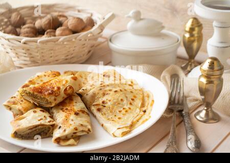Aufgerollte Crepes oder Pfannkuchen, gefüllt mit gehackter Leber auf weißem Teller auf altem Holztisch mit Vintage-Gabel und Messer Stockfoto