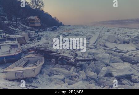 Zerstörte Boote an einem gefrorenen Donau-Fluss mit vielen Eiswürfeln und fantastischem Sonnenuntergang in der Nähe von Belgrad, Zemun, Serbien, Dunsteffekt Stockfoto