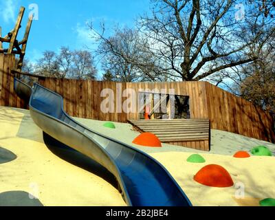 Rutsche aus Edelstahl im öffentlichen Park. Farbenfroher Sicherheitsboden aus orangefarbenem, gelbem und grünem Gummi. Fröhliche Halbkugelformen und Muster. Baumhaus. Stockfoto