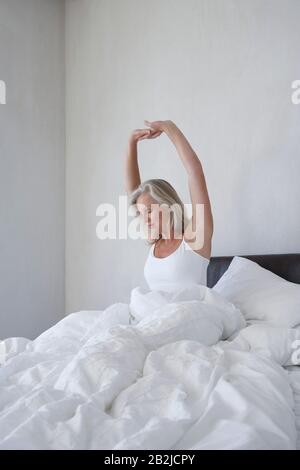Frau in Schlafanzug, die morgens im Bett unter der Decke sitzt und sich ausdehnt Stockfoto