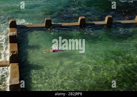 Schwimmen Sie im kühlen, grünen Meerwasserpool der Ross Jones Rockpool Coogee Bay mit Brandung, die gegen die crenulierten Umfassungsmauern, Sydney, bricht Stockfoto