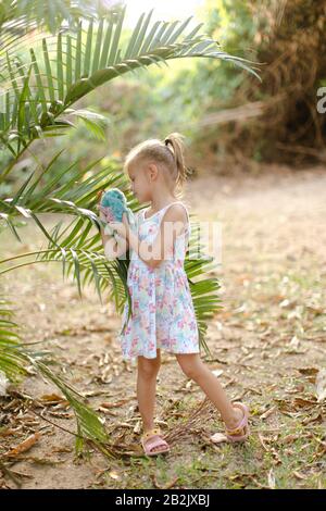 Kleines Mädchen, das mit Spielzeug spielt und in der Nähe von Palmblättern steht. Stockfoto