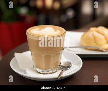 Flacher weißer Kaffee aus transparentem Glas mit weißem Schaum, hinter eclair Kuchen, Essen auf dem Tisch in einem Café Stockfoto