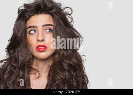 Nachdenkliche junge Frau mit roten Lippen, die seitwärts über grauem Hintergrund blickt Stockfoto