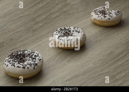 Drei Donuts mit weißer Glasur und Schokolade bestreuen auf einem Holztischhintergrund, einer davon gebissen Stockfoto